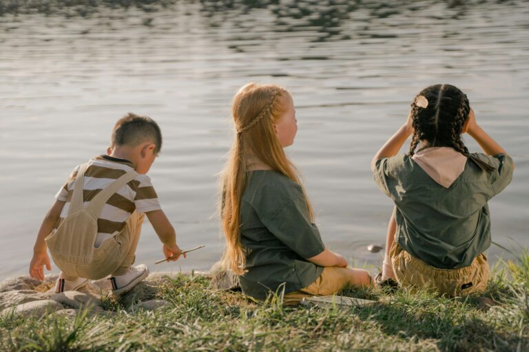 An image of three children sitting near a lake. Encourage spending time with pears helping children make friends.
