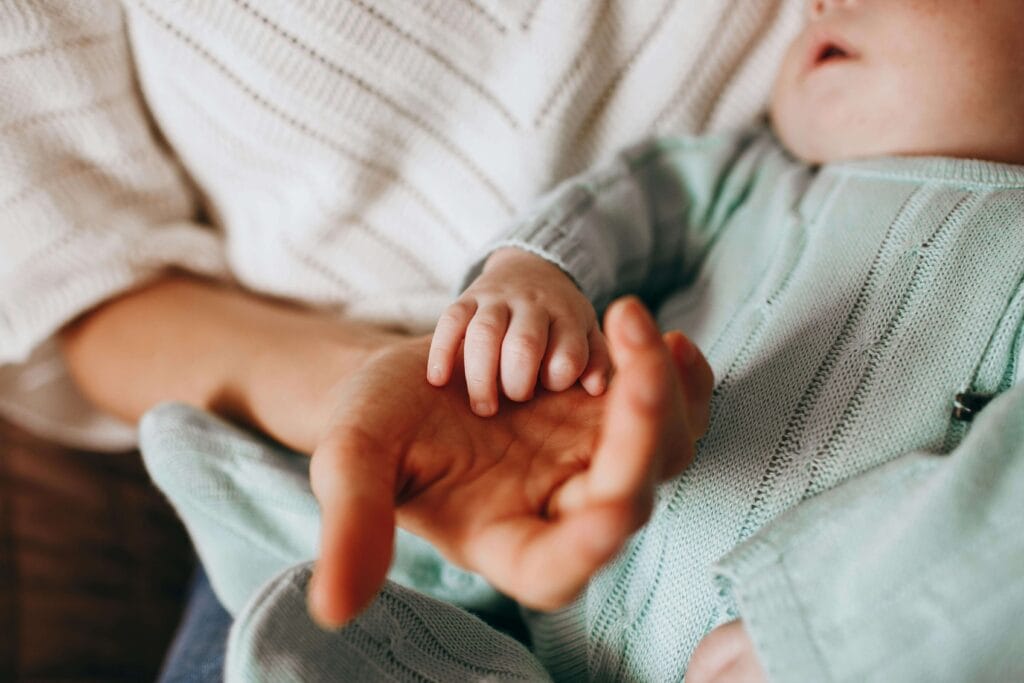 A newborn sleeps peacefully, holding her mom’s hand—a tender moment with a Funny twist: "Advice for New Parents" often means learning to sleep anywhere, anytime.